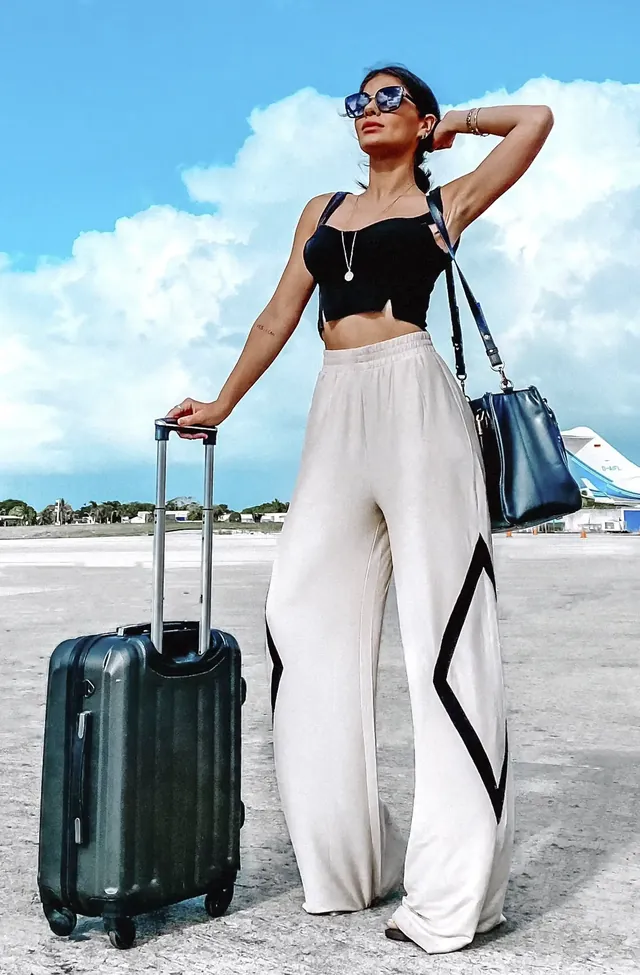 Mulher de calça pantalona com mala de viagem, de braço erguido, em pose de viagem